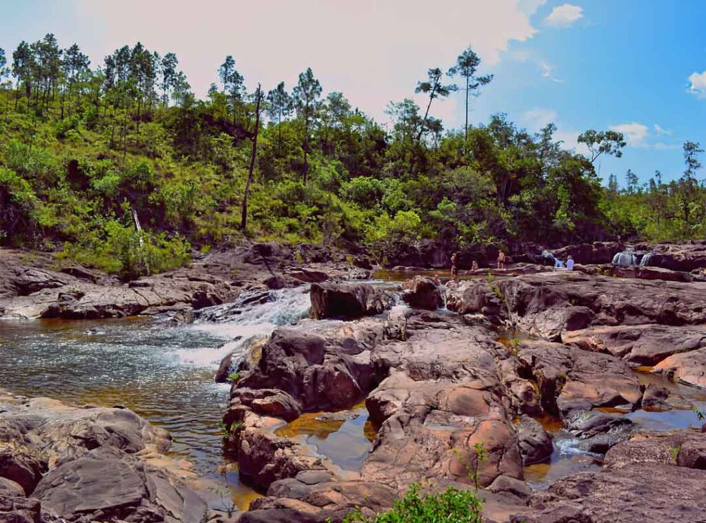 People explore a rocky riverbed with a small waterfall, surrounded by green trees under a bright blue sky on a sunny day.
