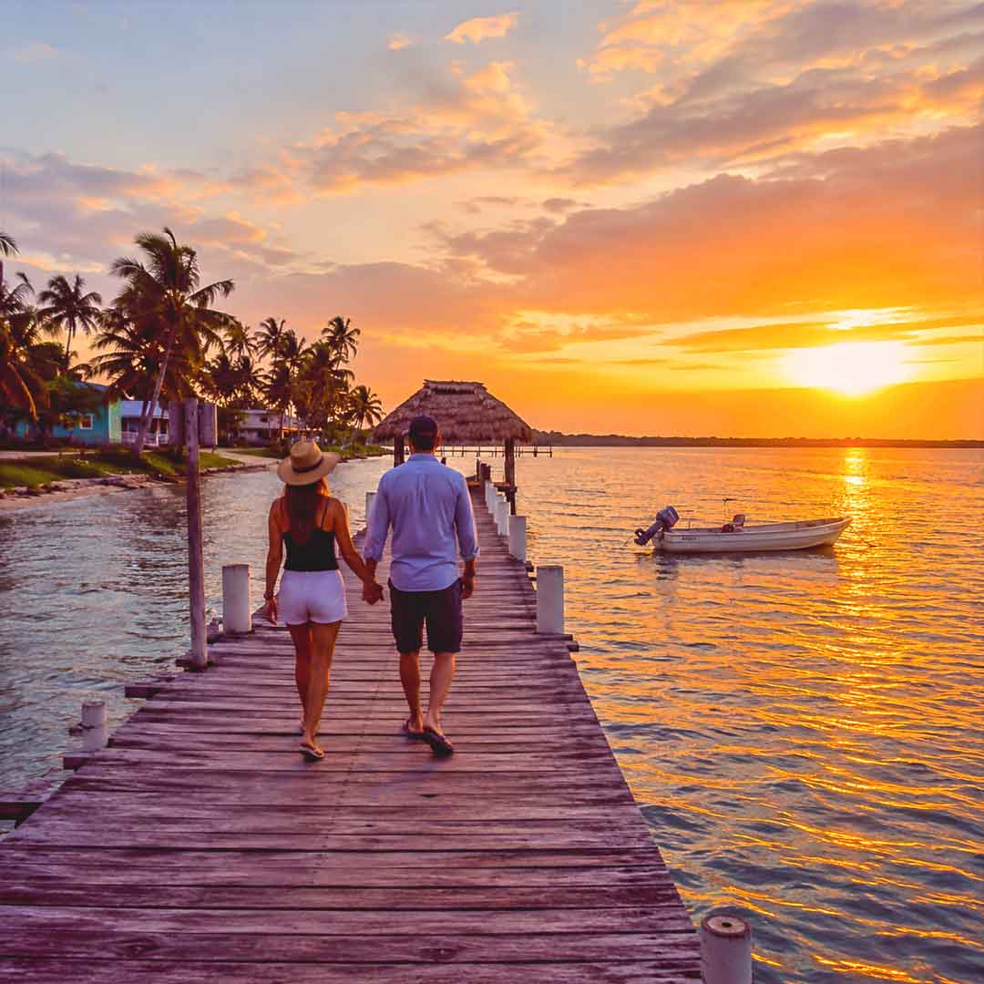 Couple walking hand in hand along a wooden pier at sunset in Belize surrounded by turquoise Caribbean waters, tropical palms, and a peaceful island atmosphere.