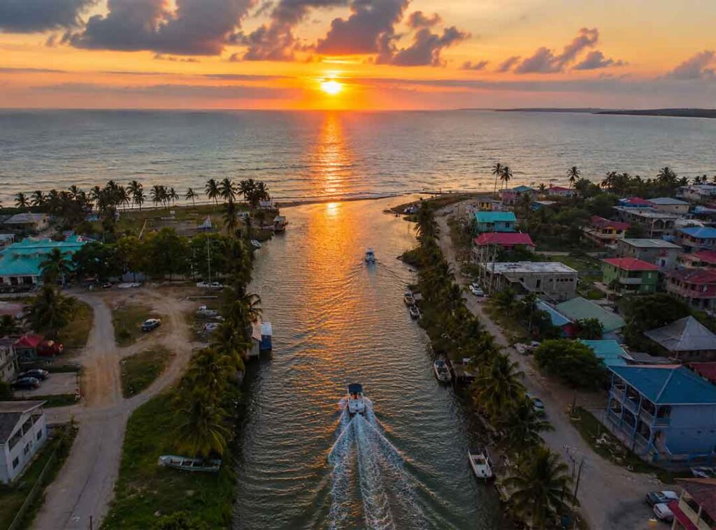 Aerial sunset view of Dangriga Belize with boats navigating a Caribbean canal lined by palm trees, colorful coastal homes, and golden reflections over the sea.