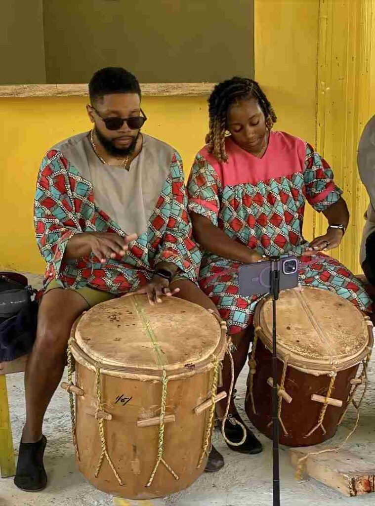 Two people in colorful matching outfits play conga drums while a smartphone on a stand records the session in front of a yellow wall.