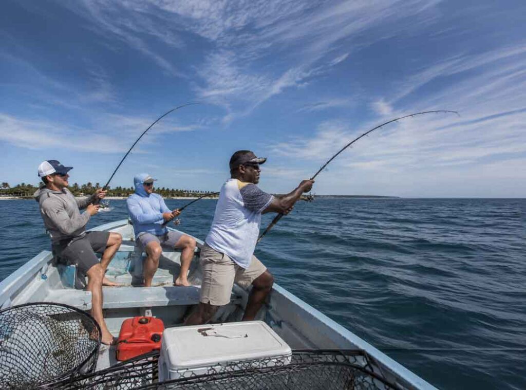 Three men fishing from a small boat on calm blue water under a clear sky.