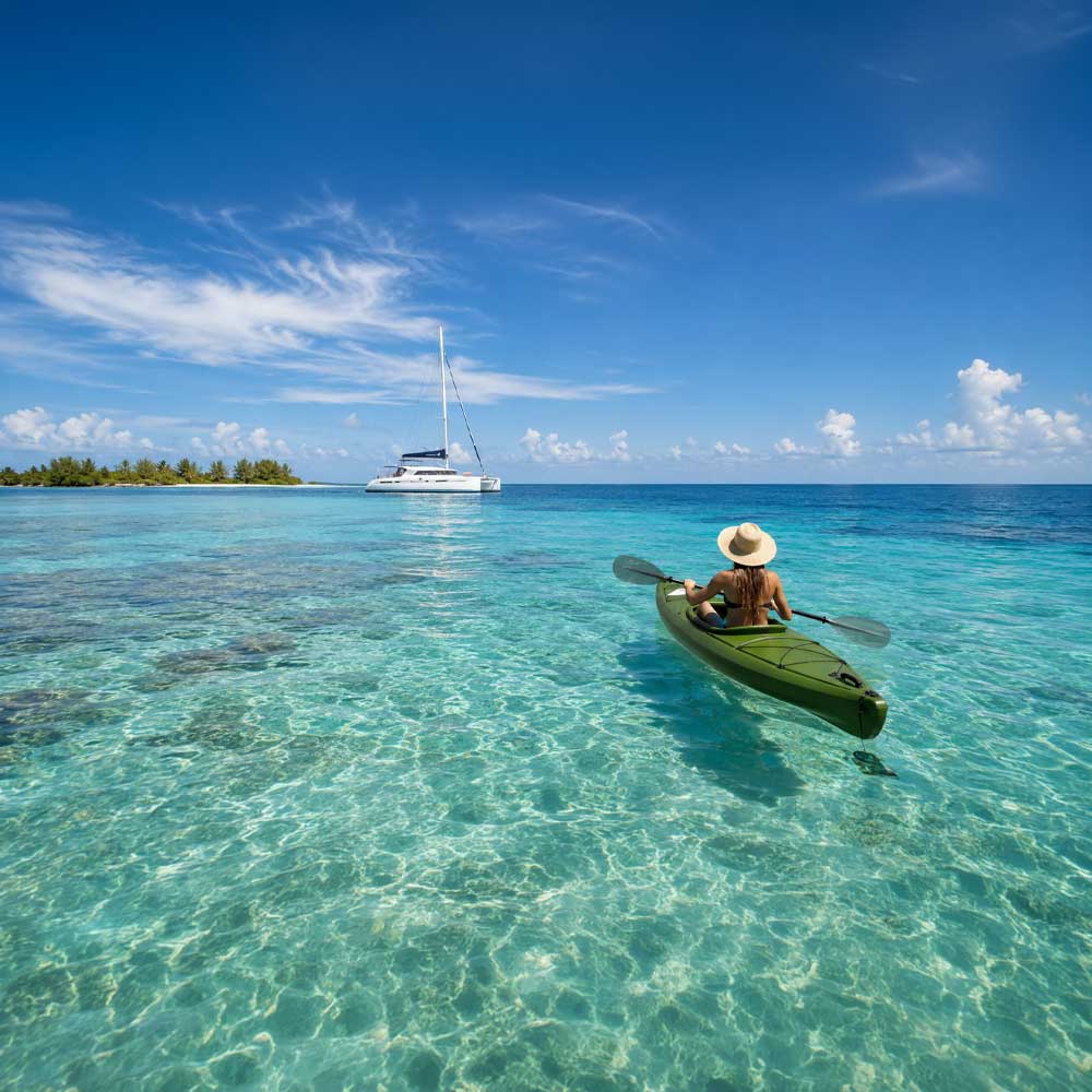 Woman kayaking in turquoise Caribbean waters during a Belize sailing vacation with a luxury catamaran anchored in the distance near the Belize Barrier Reef.