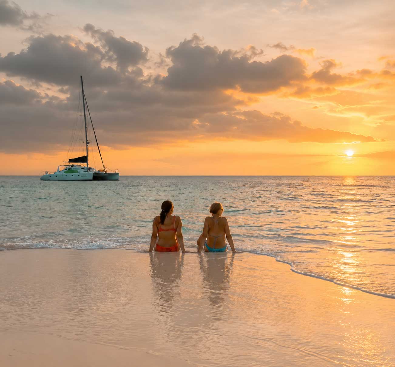 Sunset-Saliling-Belize-Vacations Two women enjoying a peaceful sunset beach moment during Belize Sailing Vacations with a luxury catamaran anchored offshore in the Caribbean Sea.