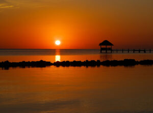 Sunset over calm water with an orange sky and a thatched gazebo on a pier to the right, silhouetted against the horizon.