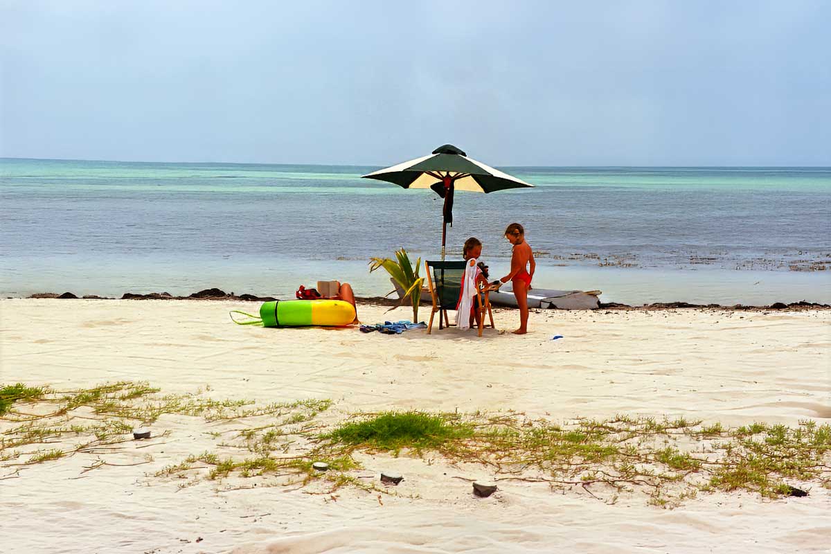 Beach-Vacation-Belize - Authentic Travel Belize Children playing under a beach umbrella on a quiet Caribbean shore during a Belize Family Vacation.