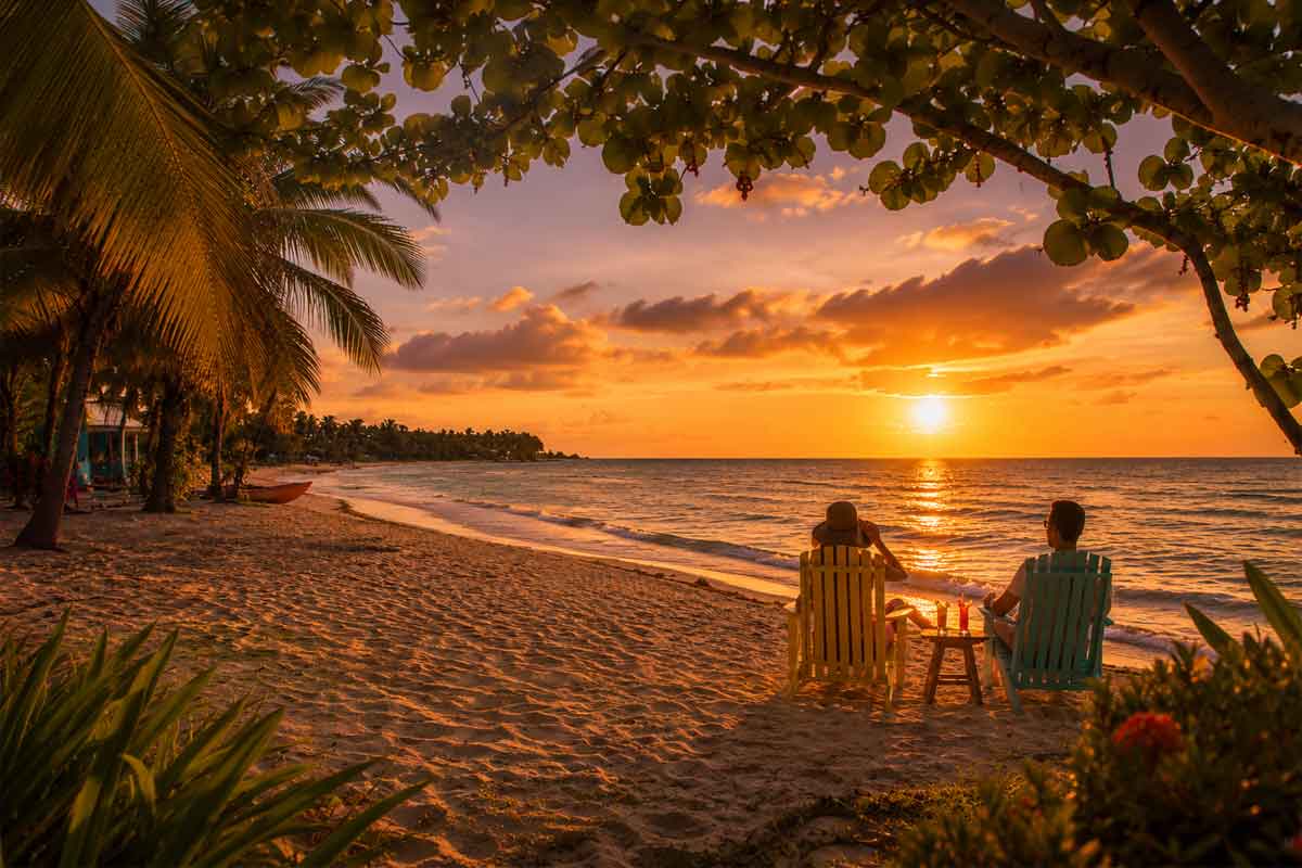 Couple seated on a Belize beach enjoying tropical drinks at Caribbean sunset, showcasing romantic Belize travel styles for a relaxed coastal escape.
