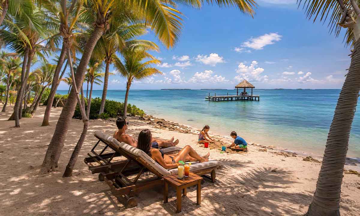 Coast-&-Relaxation-Family-Journey Belize Family Vacations with parents relaxing in beach chairs while children play in the sand beside clear turquoise water and the Belize Barrier Reef.