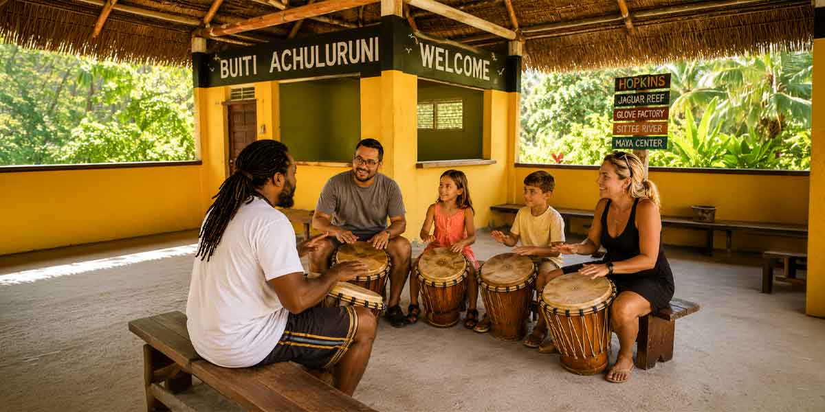 Cultural-Adventure-Belize Group of people (adults and children) sitting on benches in a yellow open-air shelter, playing djembes together in a drumming circle.