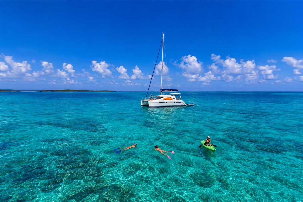 Mother kayaking beside her child snorkeling in turquoise Belize waters, with a catamaran in the background, highlighting family-friendly Belize travel styles.